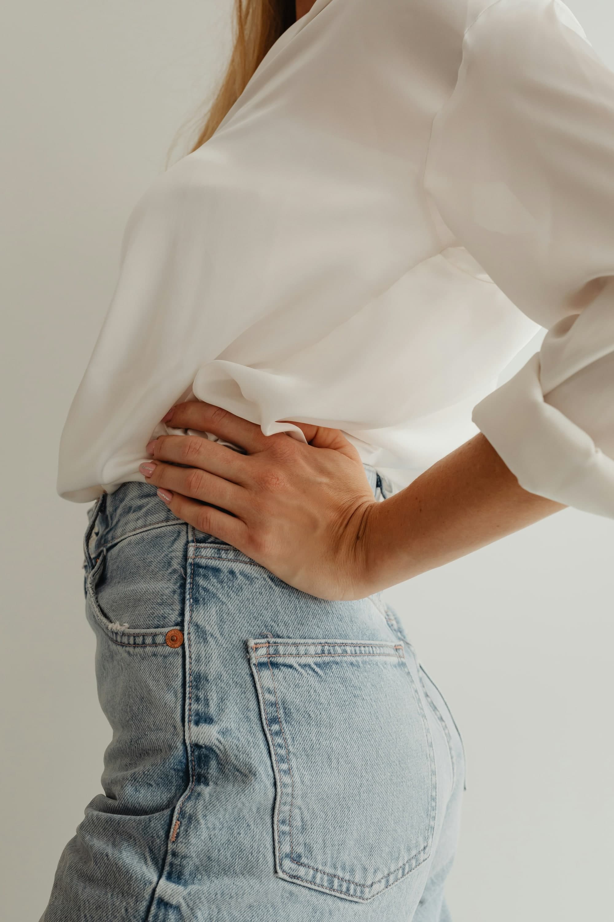 A white blouse tucked into worn high-waisted denim, a hand smoothing it at the hip.